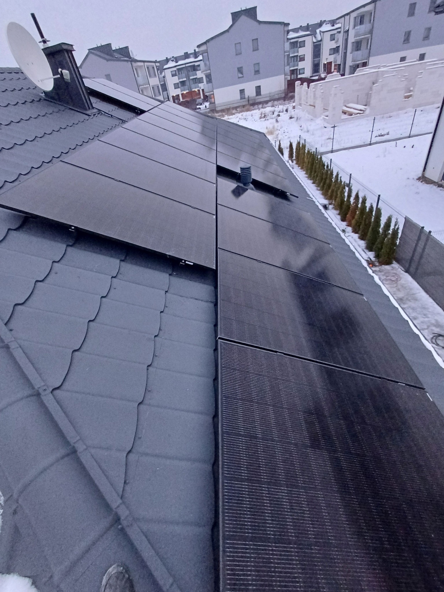 Czarne panele słoneczne na szarym dachu skośnym pokrytym śniegiem, w tle budynki mieszkalne i częściowo zasypany śniegiem teren.