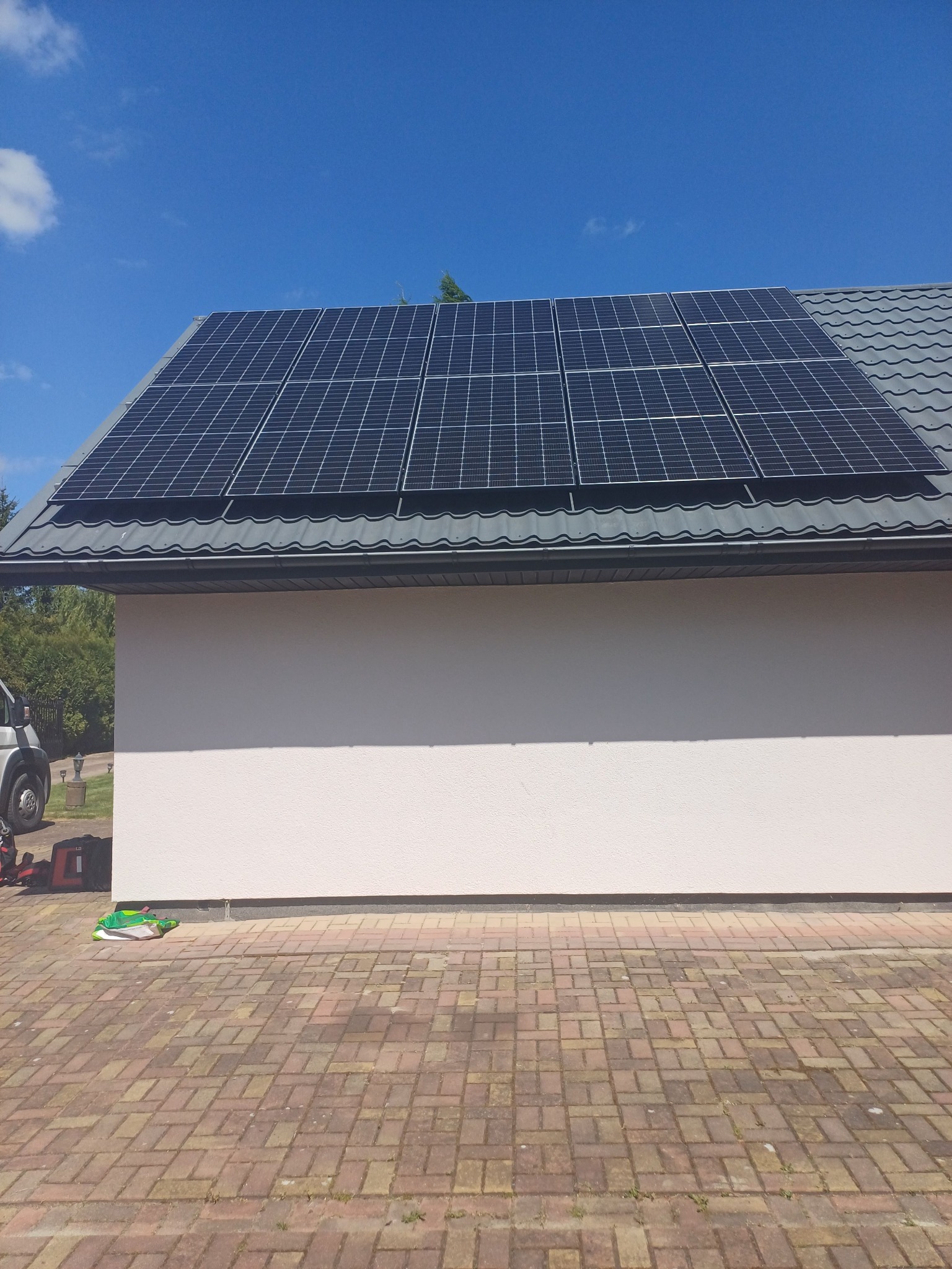 Panele słoneczne na ciemnoszarym dachu dwuspadowym budynku z białą elewacją, widoczny fragment samochodu dostawczego i kostka brukowa na podjeździe.
