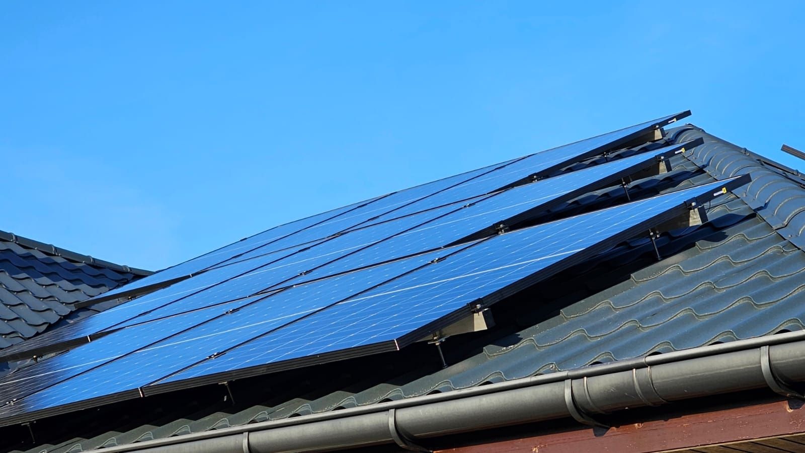 Instalacja paneli słonecznych na dachu z ciemnej dachówki ceramicznej, widoczne mocowania i rynna spustowa na tle błękitnego nieba.
