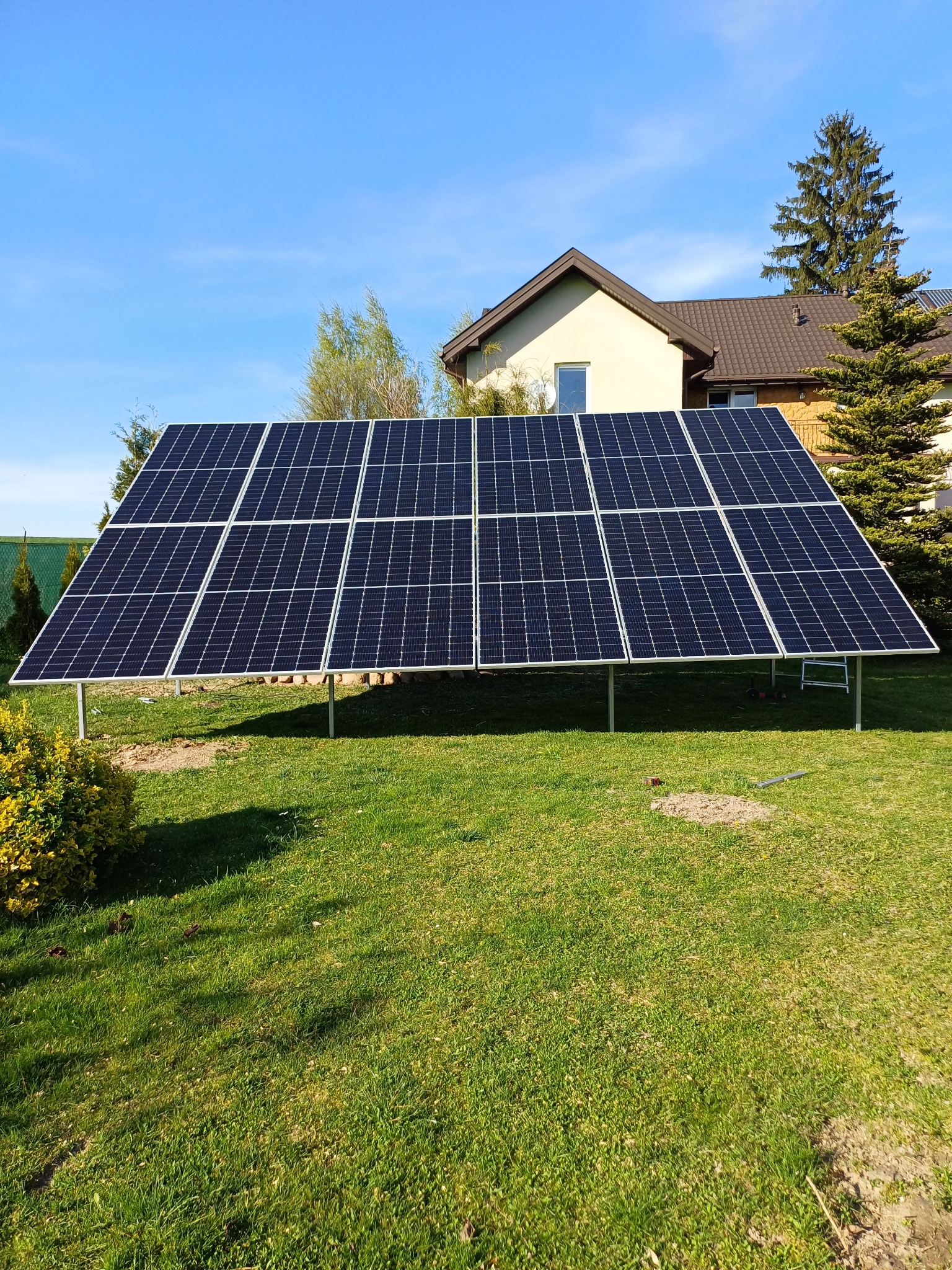 Instalacja fotowoltaiczna naziemna na metalowej konstrukcji, widok z boku na tle domu jednorodzinnego i zielonego trawnika w słoneczny dzień.