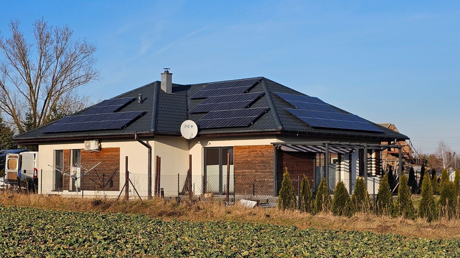 Dom jednorodzinny z ciemną dachówką i panelami słonecznymi na dachu, widoczna antena satelitarna, jasna elewacja z elementami drewnianymi, otoczony polem i ogrodzeniem.