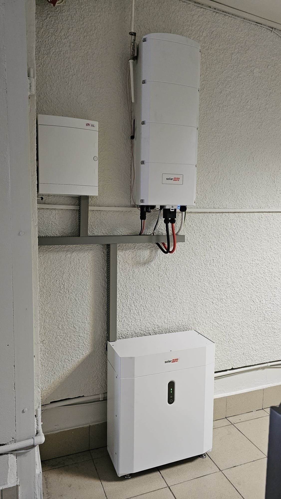 Biała skrzynka elektryczna ETI na ścianie, powyżej biały inwerter SolarEdge zamontowany na szarej metalowej konstrukcji, a na podłodze biały magazyn energii SolarEdge z diodami LED, na tle ściany...