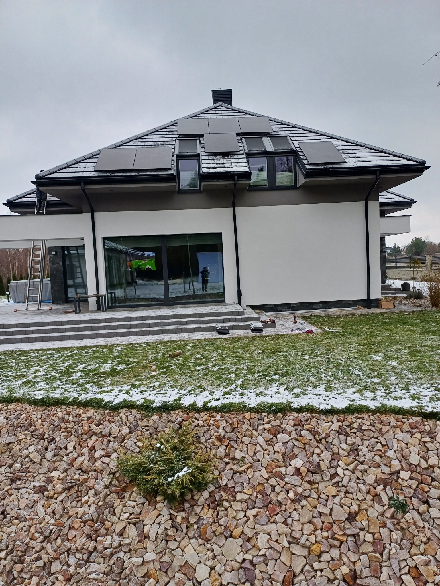 Nowoczesny dom jednorodzinny z panelami słonecznymi na dachu pokrytym dachówką, widoczne okna dachowe, trawnik z resztkami śniegu, kamienne umocnienie skarpy i drabina oparta o ścianę budynku.