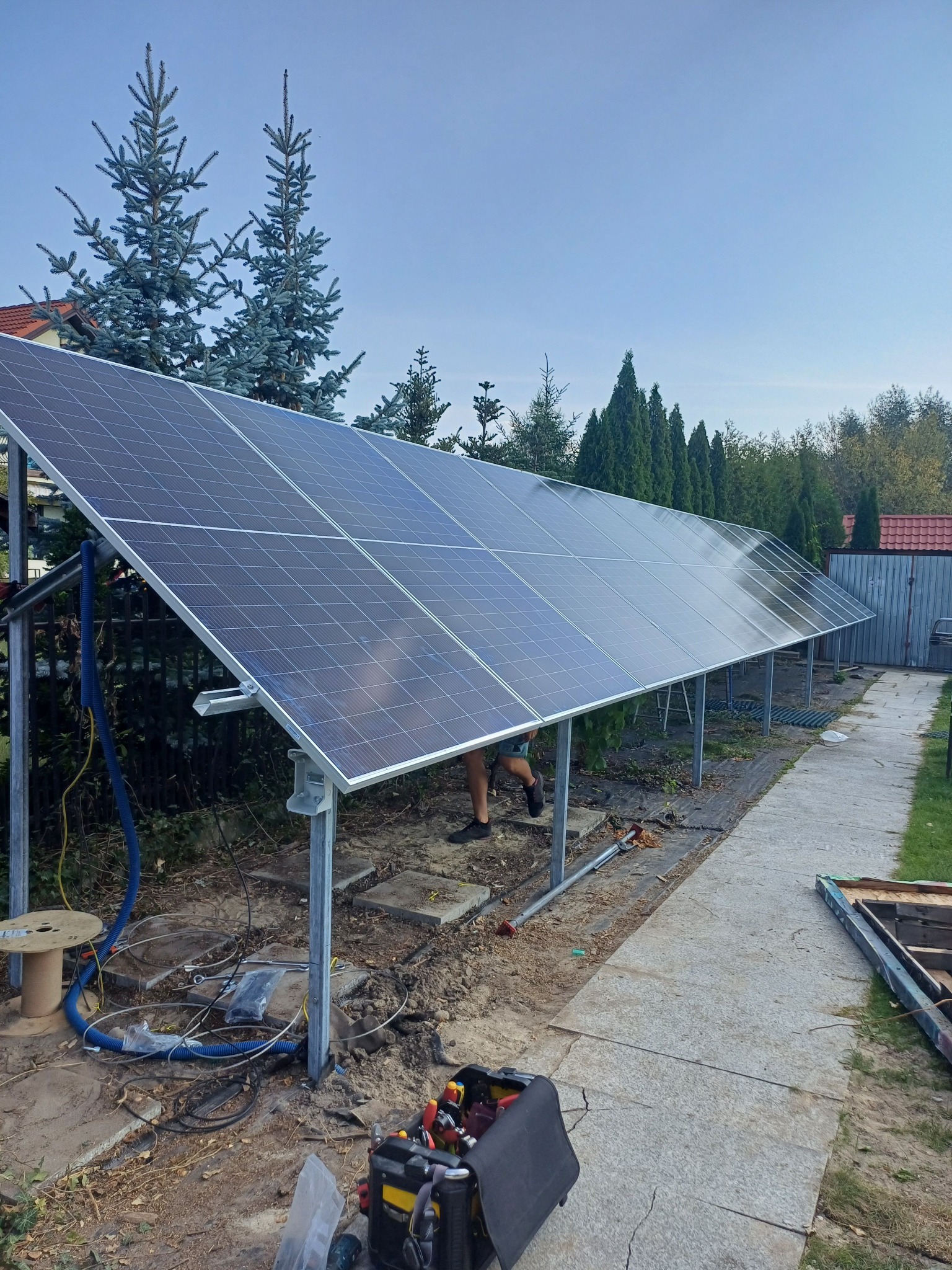 Długi rząd paneli na metalowej konstrukcji w ogrodzie, widoczne narzędzia i kable sugerują montaż w toku, błękitne świerki w tle.