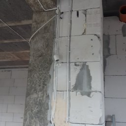 EneGo Michał Pilarski - Wewnętrzne kable elektryczne w białych rurkach przymocowane do surowej ściany z pustaków, widoczne puszki pod gniazdka. Etap prac instalacyjnych.