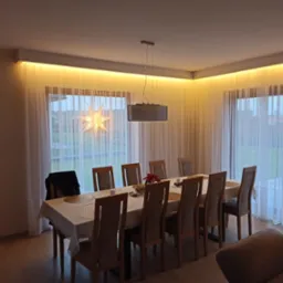 Jadalnia z oświetleniem LED podsufitowym i lampą wiszącą. Widoczne okna z firanami i dekoracją w kształcie gwiazdy. Ciepłe światło tworzy przytulną atmosferę.