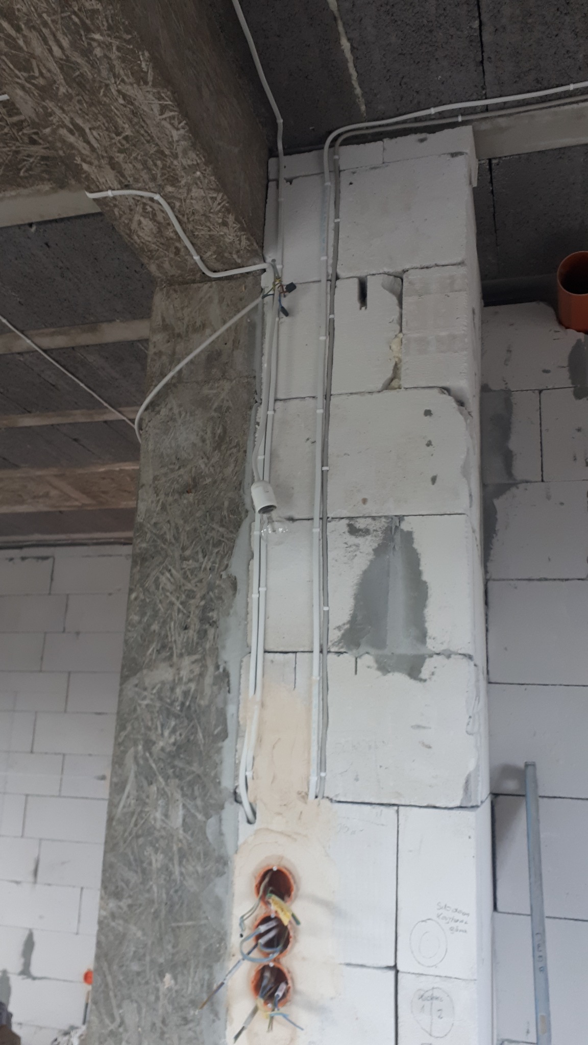 Wewnętrzne kable elektryczne w białych rurkach przymocowane do surowej ściany z pustaków, widoczne puszki pod gniazdka. Etap prac instalacyjnych.
