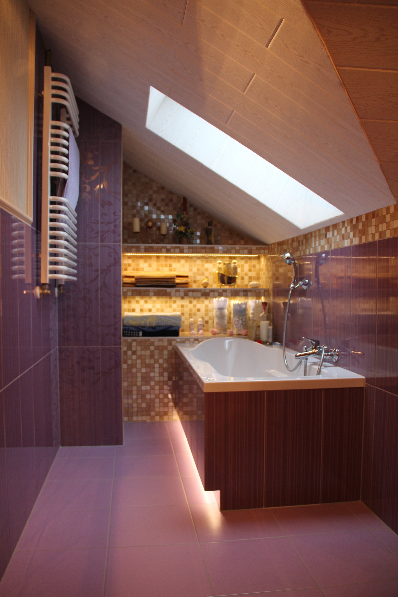 Łazienka na poddaszu z oknem dachowym, oświetleniem LED pod wanną i na półkach. Grzejnik drabinkowy, mozaika i fioletowe płytki tworzą spójną całość.