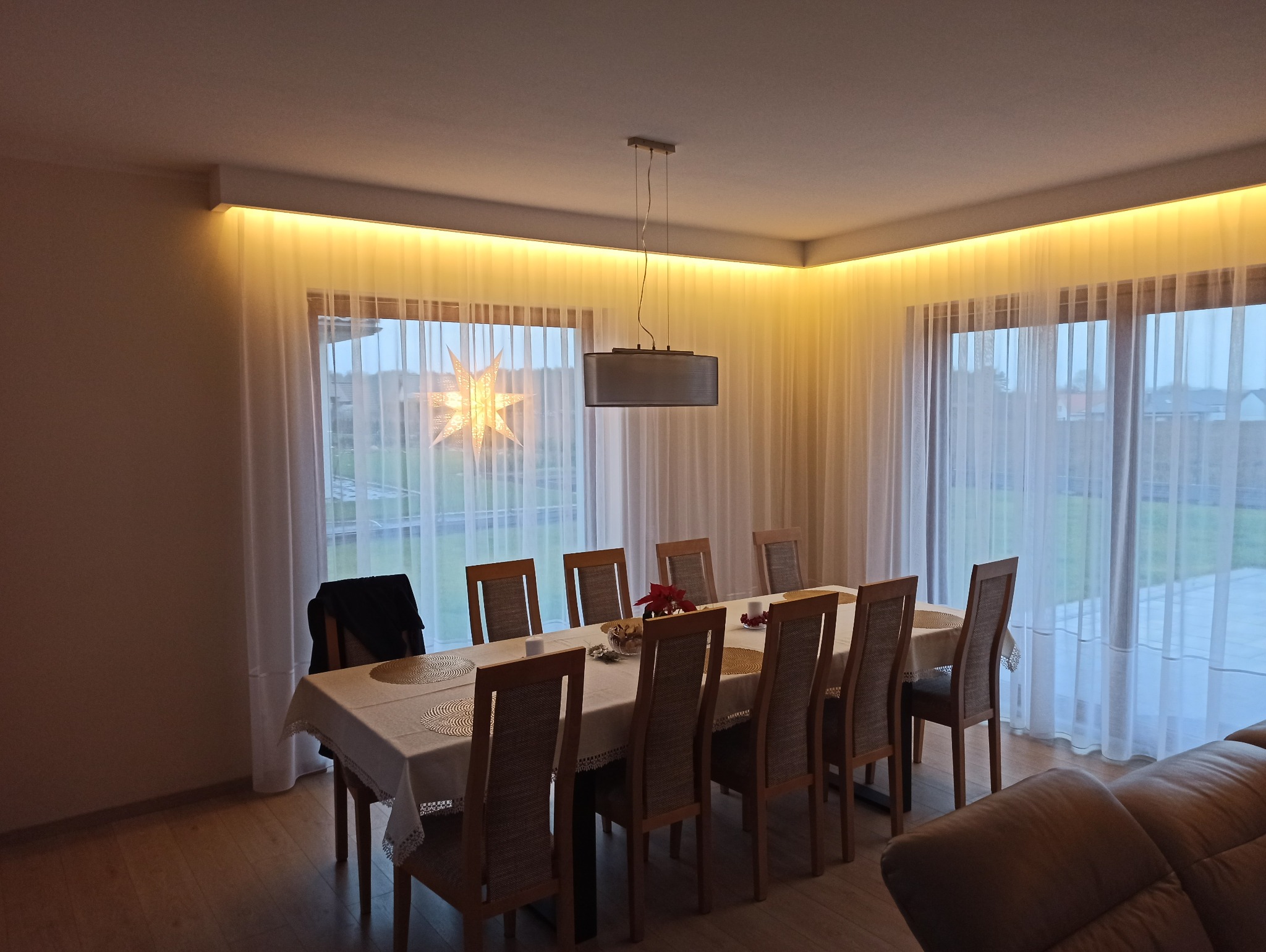 Jadalnia z oświetleniem LED podsufitowym i lampą wiszącą. Widoczne okna z firanami i dekoracją w kształcie gwiazdy. Ciepłe światło tworzy przytulną atmosferę.