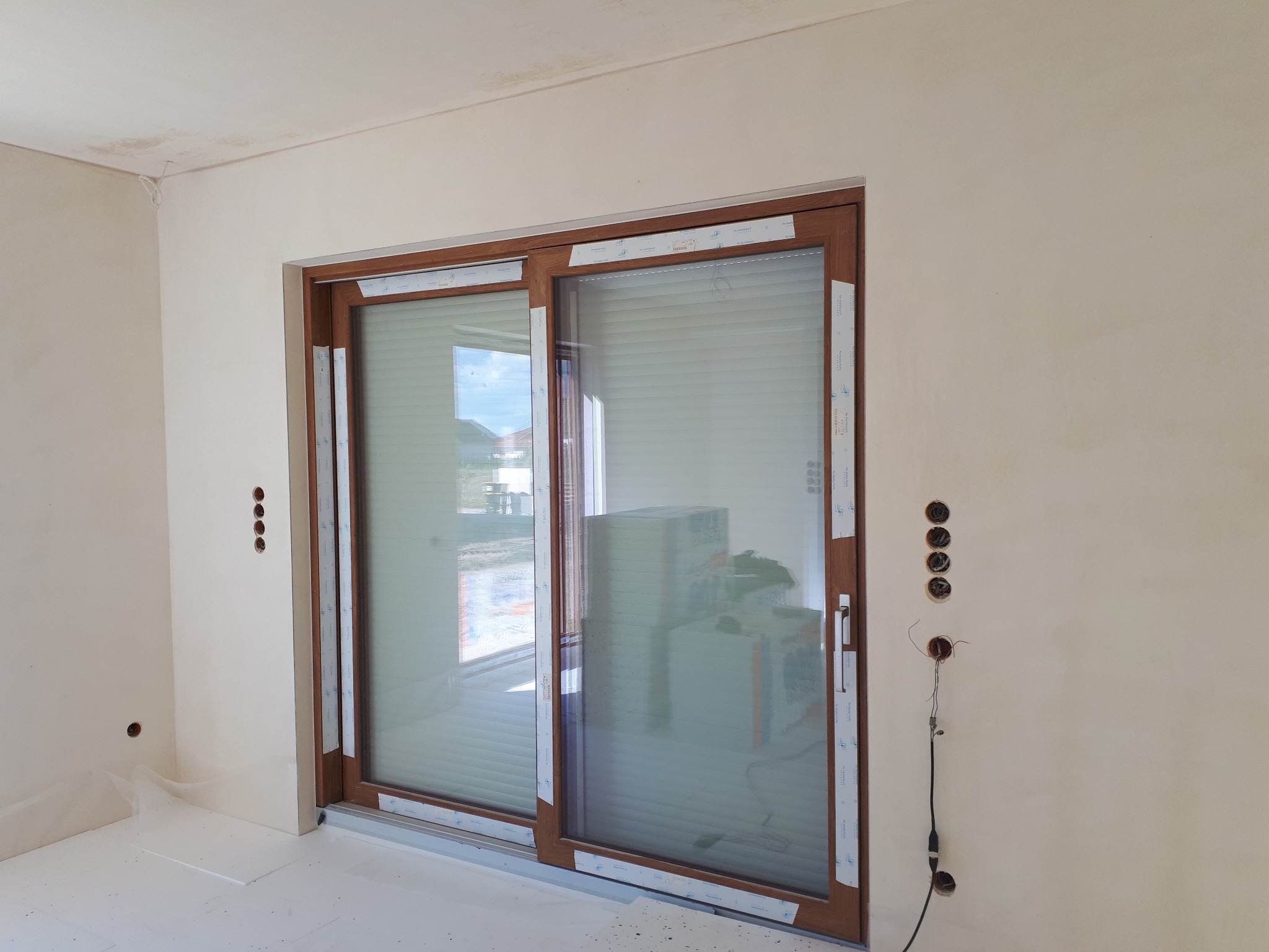 Surowy stan wnętrza: przesuwne okno balkonowe z brązową ramą, ściany w trakcie wykończenia, widoczne puszki elektryczne bez osprzętu i wystające przewody.