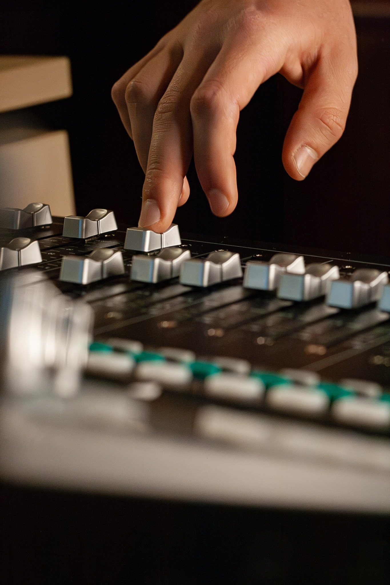 Dłoń reguluje suwak na profesjonalnym mikserze audio z podświetlanymi przyciskami, widoczne detale skóry i odbicia światła na metalowych elementach.