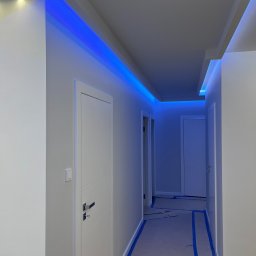 Wiwian sp. z o.o. - Biały korytarz z niebieskim oświetleniem LED na suficie, widoczne drzwi i zabezpieczona podłoga taśmą malarską. Nowoczesne wykończenie wnętrza.