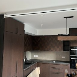 Wiwian sp. z o.o. - Nowoczesna kuchnia z ciemnego drewna z geometrycznym wzorem na szafkach, szarym blatem i heksagonalną podłogą w trakcie wykończenia. Widoczne kartony i elementy montażowe.