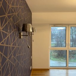 Wiwian sp. z o.o. - Pokój po remoncie: wzorzysta tapeta w odcieniach granatu i złota, drewniana podłoga, nowoczesne kinkiety i okno z widokiem na zewnątrz. Minimalistyczny design.