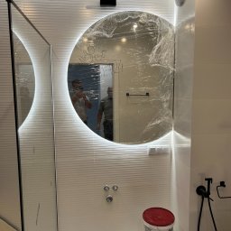 Wiwian sp. z o.o. - Nowoczesna łazienka w trakcie wykończenia: okrągłe lustro LED zabezpieczone folią, białe płytki z poziomymi paskami, widoczne elementy instalacji i pojemniki z farbą.