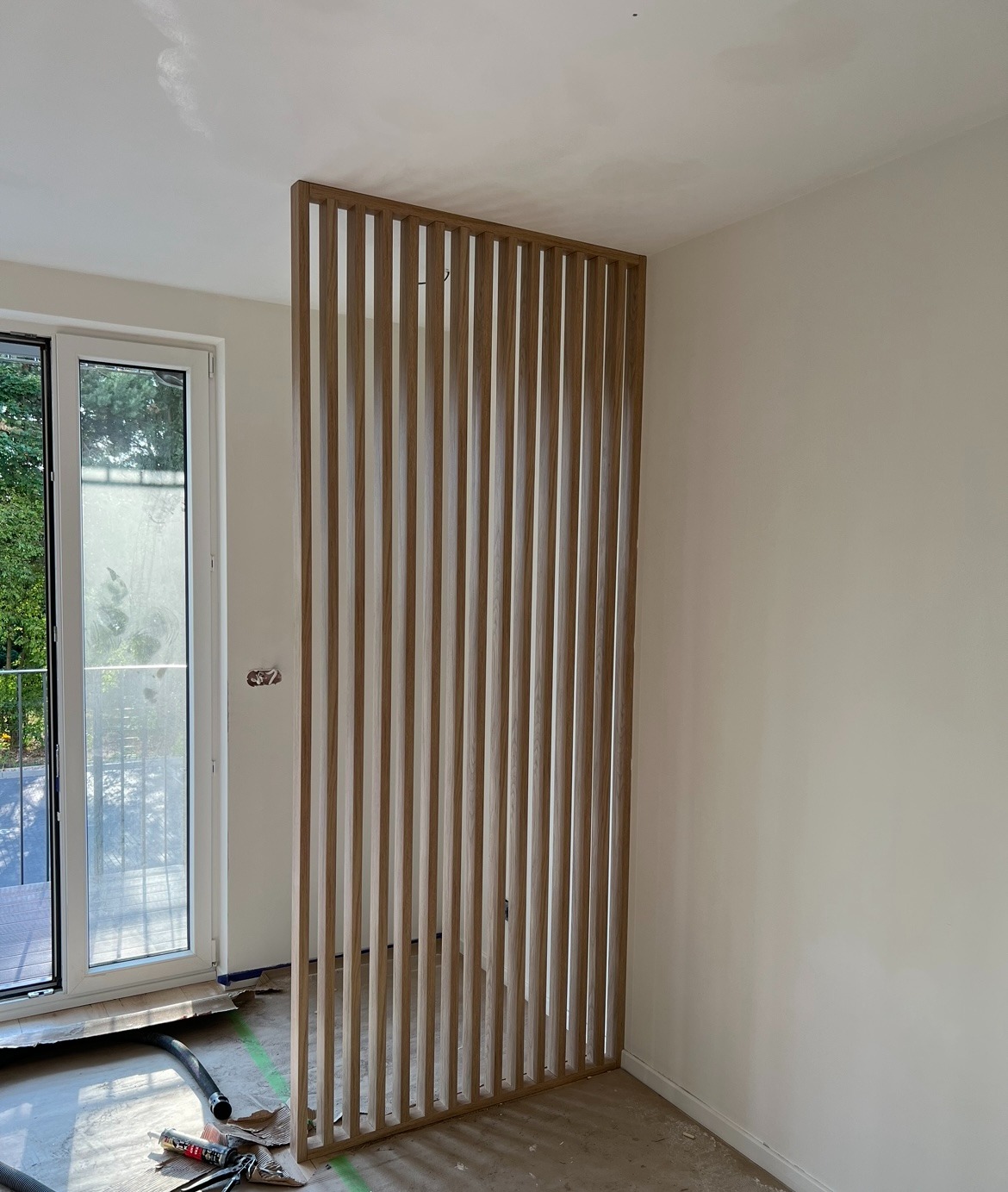 Drewniana ażurowa ścianka działowa z pionowych listew, oddzielająca przestrzeń w jasnym wnętrzu. Widoczne okno balkonowe i surowy stan podłogi.