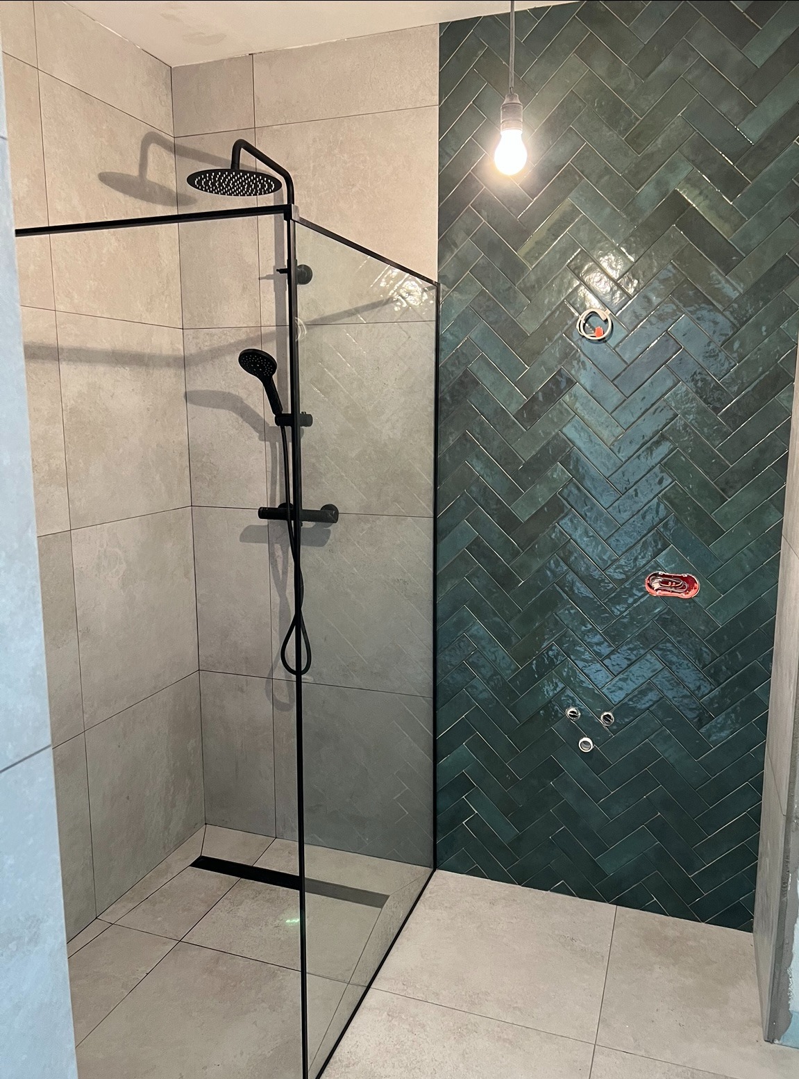 Nowoczesna kabina prysznicowa z czarną armaturą i zieloną ścianą z płytek ułożonych w jodełkę. Minimalistyczny design, surowy klimat, widoczne przyłącza.