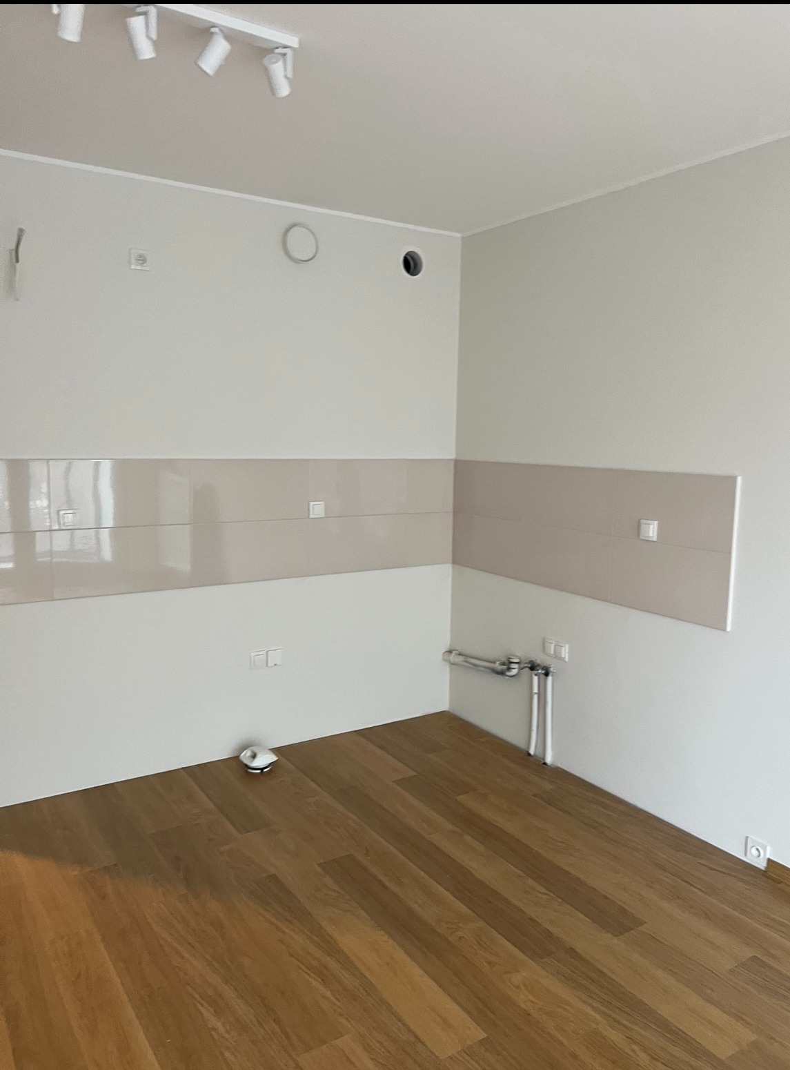 Jasna kuchnia po remoncie: drewniana podłoga, białe ściany z beżowymi płytkami, widoczne rury i instalacja wodna. Nowoczesne oświetlenie sufitowe.