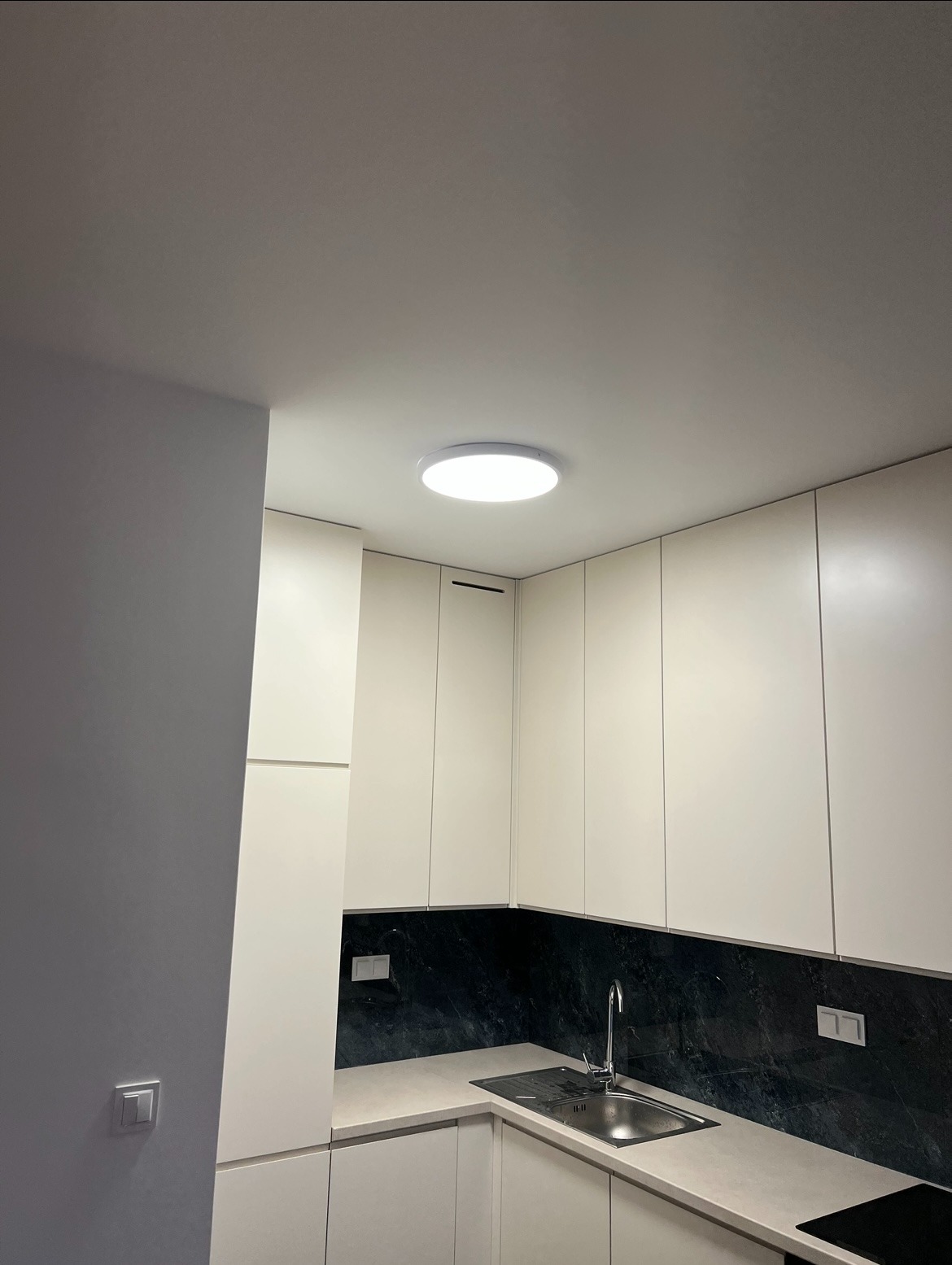 Nowoczesna kuchnia z białymi szafkami, czarnym blatem i okrągłą lampą sufitową. Minimalistyczny design, widok na narożnik blatu i zlew.