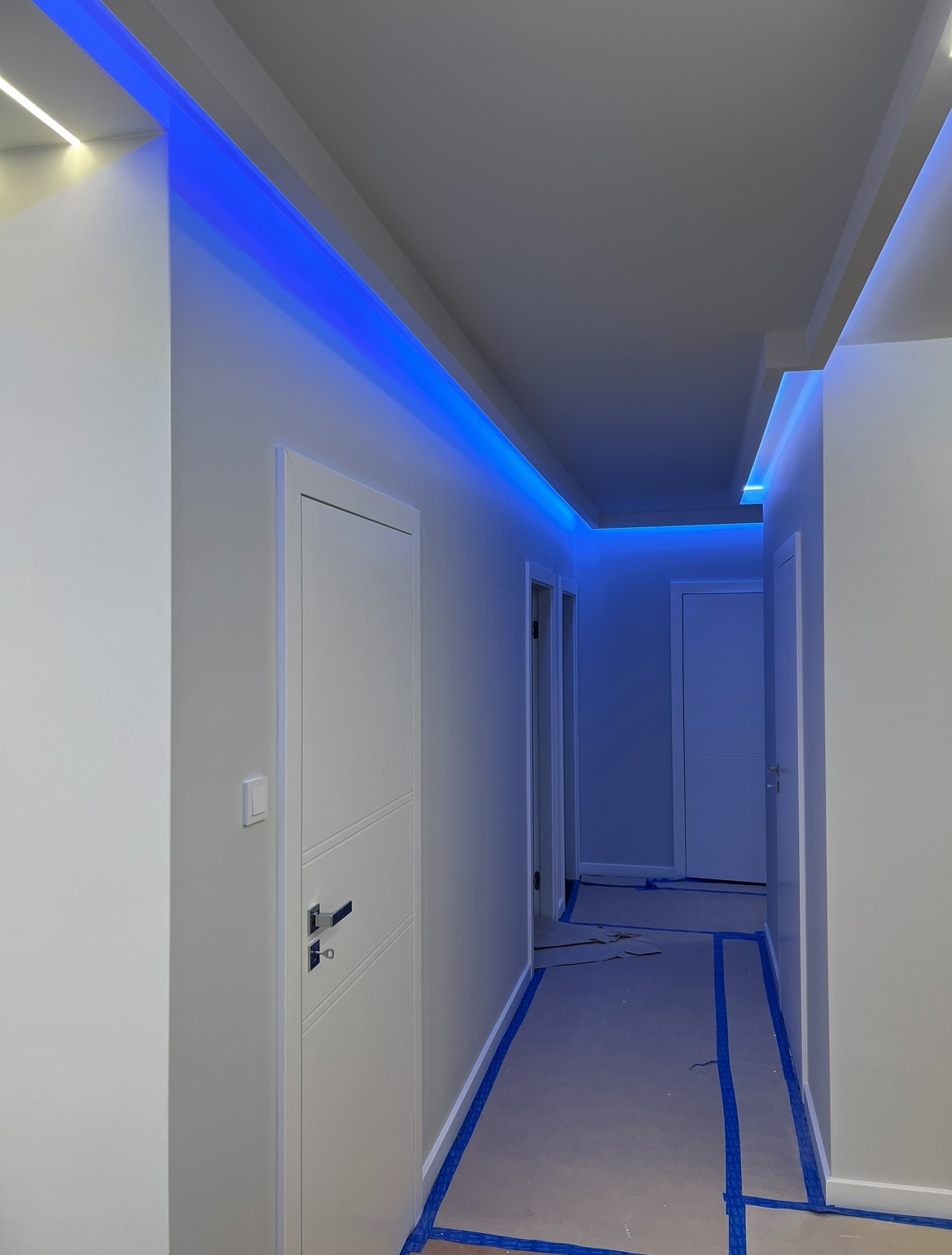 Biały korytarz z niebieskim oświetleniem LED na suficie, widoczne drzwi i zabezpieczona podłoga taśmą malarską. Nowoczesne wykończenie wnętrza.