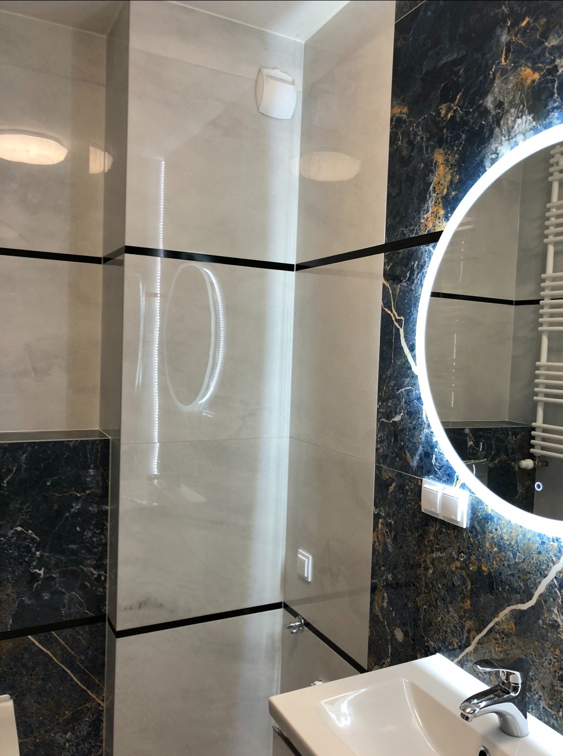 Wykończona łazienka z marmurowymi płytkami w odcieniach bieli i granatu, z okrągłym lustrem LED i grzejnikiem drabinkowym. Widoczna umywalka z baterią.