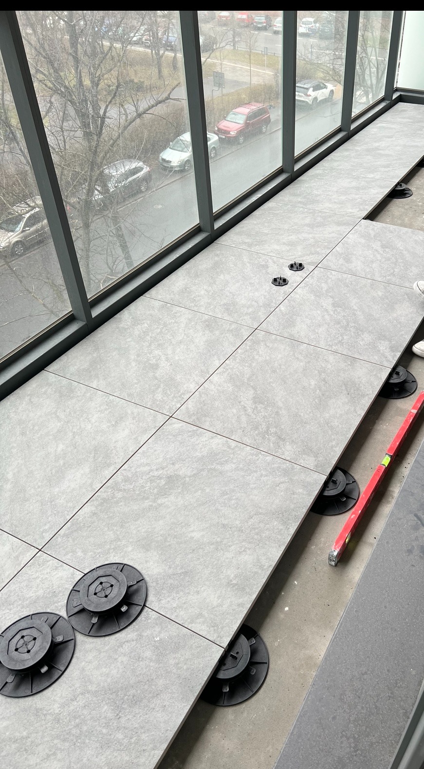 Montaż szarej, kwadratowej terakoty na regulowanych wspornikach na balkonie z widokiem na ulicę. Poziomica wskazuje na precyzję układania.