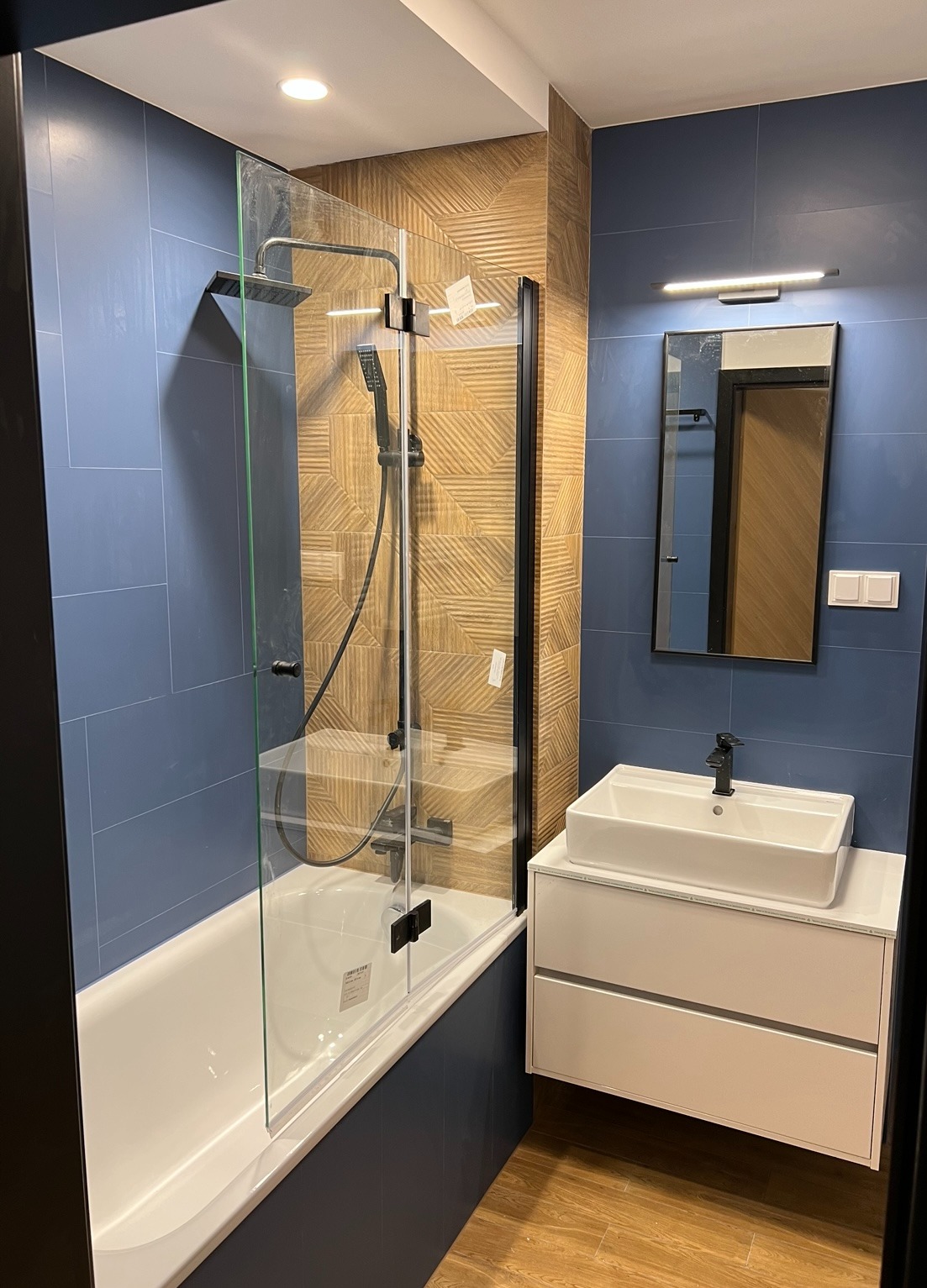 Niebieska łazienka z kabiną prysznicową typu walk-in, umywalką nablatową i lustrem z oświetleniem. Na ścianie dekoracyjna mozaika w odcieniach brązu.