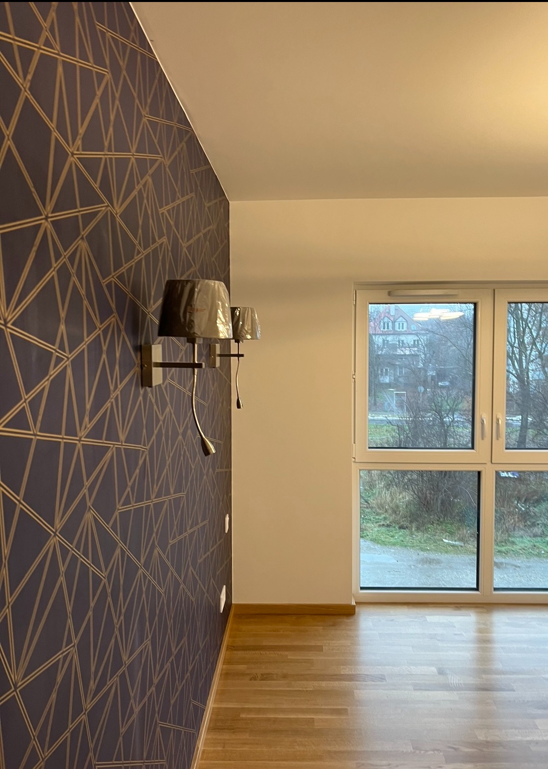 Pokój po remoncie: wzorzysta tapeta w odcieniach granatu i złota, drewniana podłoga, nowoczesne kinkiety i okno z widokiem na zewnątrz. Minimalistyczny design.