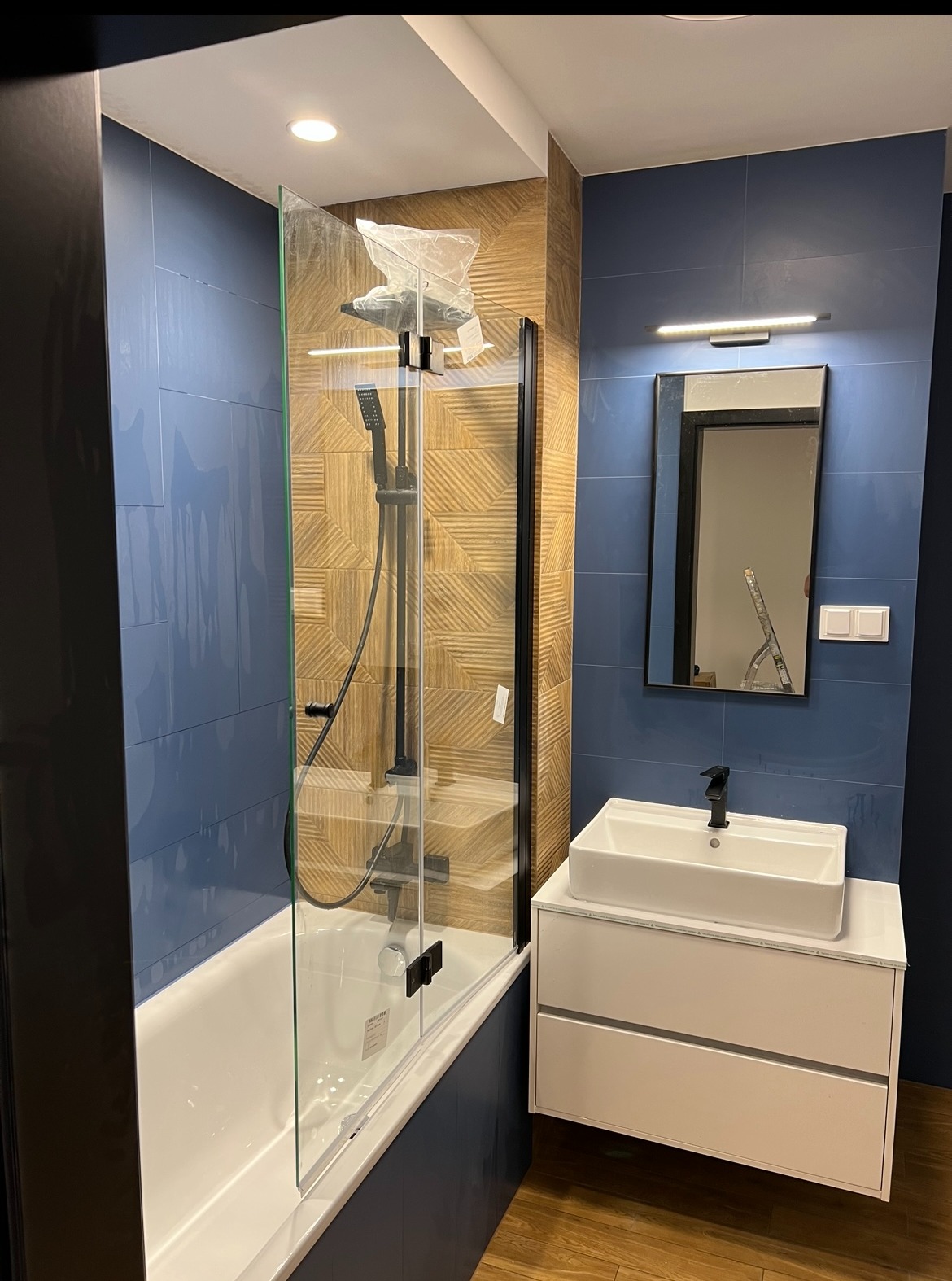 Nowoczesna łazienka z kabiną prysznicową typu walk-in, umywalką nablatową i lustrem. Ściany wyłożone niebieskimi płytkami, a strefa prysznica drewnopodobnymi panelami.