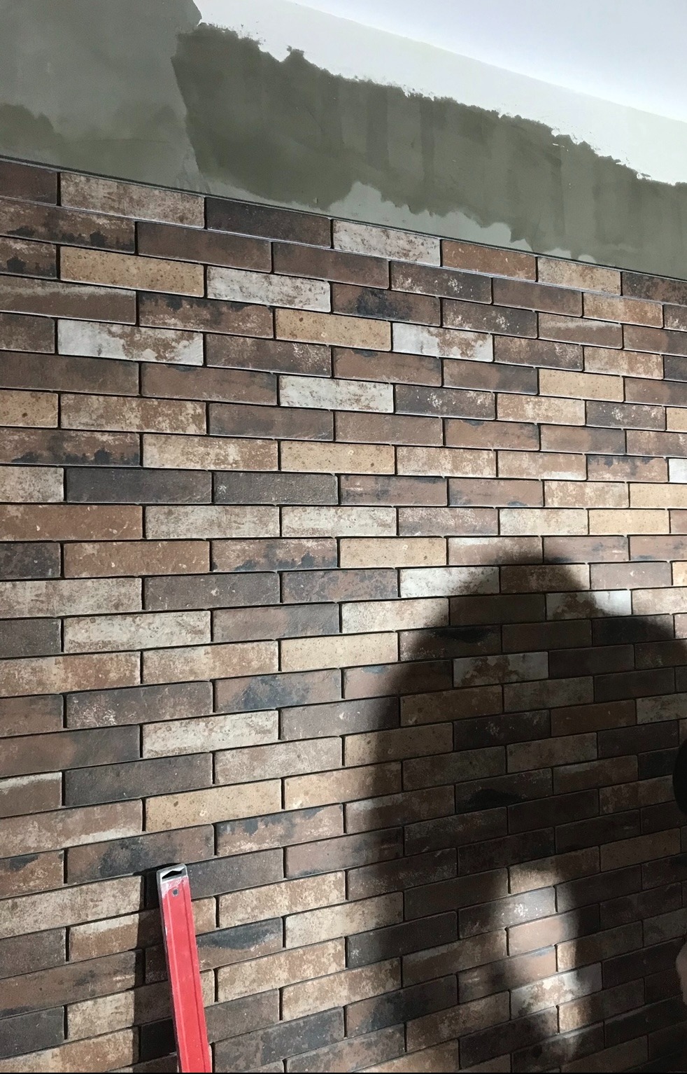 Ściana wyłożona brązowo-beżową cegłą klinkierową, z widocznym poziomowaniem i fragmentem sufitu z niedokończonym wykończeniem gładzią.