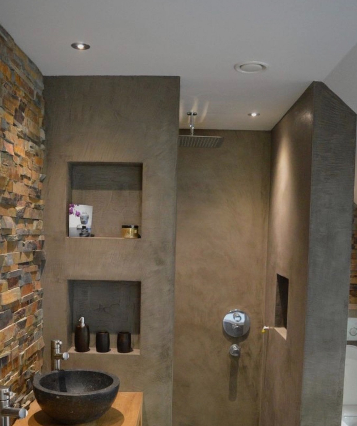 Nowoczesna łazienka z kamienną ścianą, umywalką z czarnego kamienia i prysznicem typu deszczownica. Ściany wykończone w odcieniach szarości z wnękami na akcesoria.