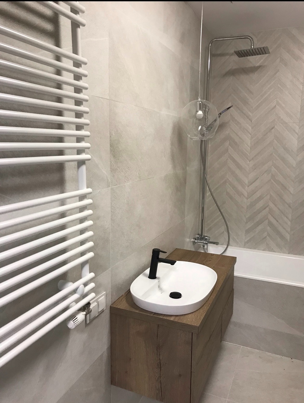Nowoczesna łazienka z owalną umywalką na drewnianej szafce, grzejnikiem drabinkowym i deszczownicą. Ściany wyłożone płytkami w jodełkę, neutralna kolorystyka.