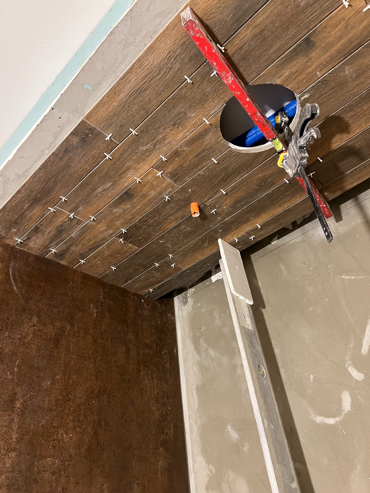 Układanie płytek imitujących drewno na suficie w łazience. Widoczne krzyżyki dystansowe, poziomica i otwór na oświetlenie. Brązowa ściana z efektem rdzy w tle.