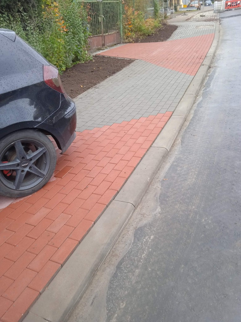 Ułożona z czerwono-szarej kostki nowa nawierzchnia chodnika, częściowo zajęta przez zaparkowany samochód, z widocznym obrzeżem i fragmentem ulicy.