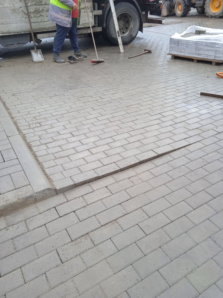 Układanie szarej kostki brukowej na podłożu, widoczne narzędzia pracy, paleta z kostką w tle, pracownik w odblaskowej kamizelce zamiata powierzchnię.