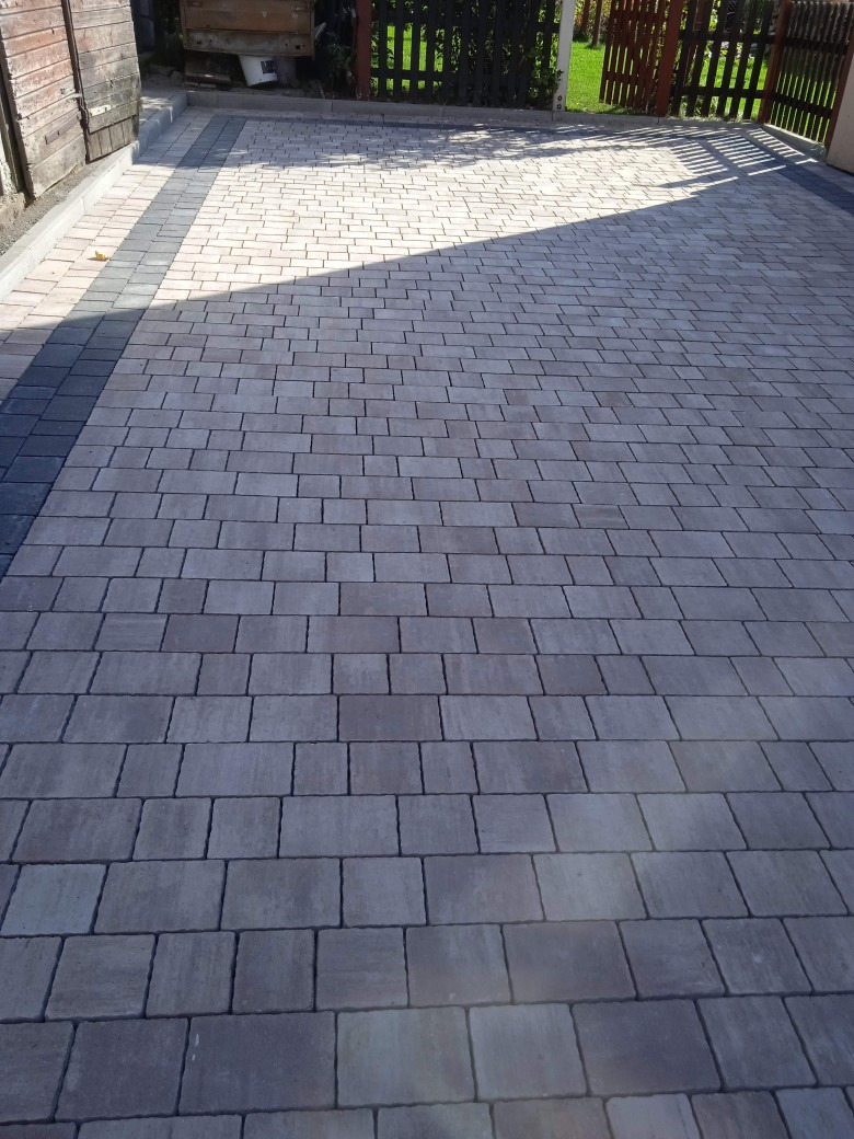 Ułożona szara kostka brukowa na podjeździe, z ciemniejszym pasem kostki po lewej stronie i drewnianym ogrodzeniem w tle.