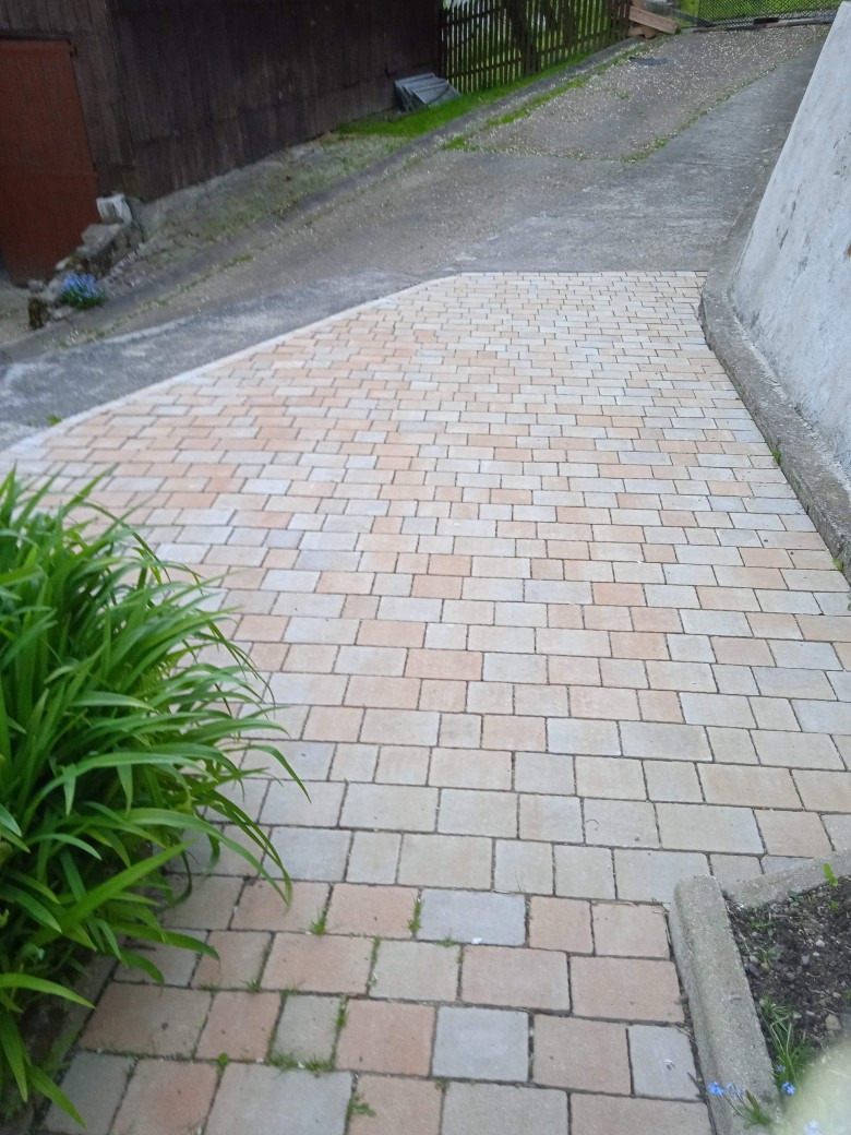 Ułożona z beżowo-różowej kostki brukowej ścieżka prowadząca do góry, z zieloną roślinnością po lewej stronie i betonowym obrzeżem po prawej, sąsiadująca z asfaltową drogą.