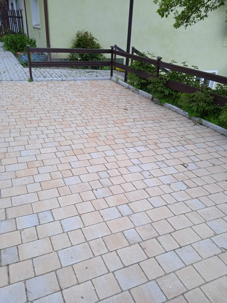 Jasna kostka brukowa ułożona na podjeździe przed domem z brązowym ogrodzeniem, widoczne rośliny i kwiaty.