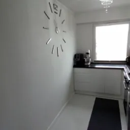 Minimalistyczna kuchnia z białymi ścianami, podłogą i meblami, kontrastującymi z czarnym blatem i dywanem, oświetlona kryształowym żyrandolem, z dużym zegarem ściennym.