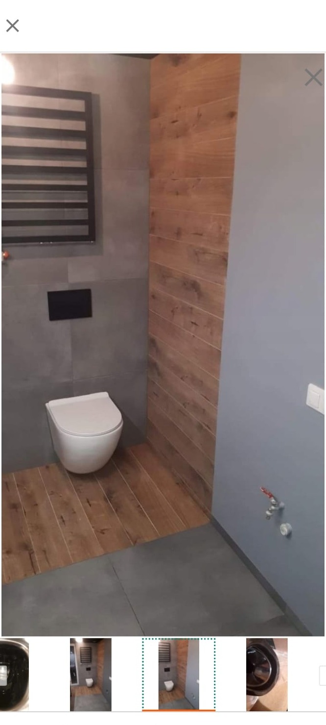 Nowoczesna toaleta z szarymi płytkami, drewnopodobną ścianą i podłogą, wisząca muszla klozetowa oraz grzejnik drabinkowy.