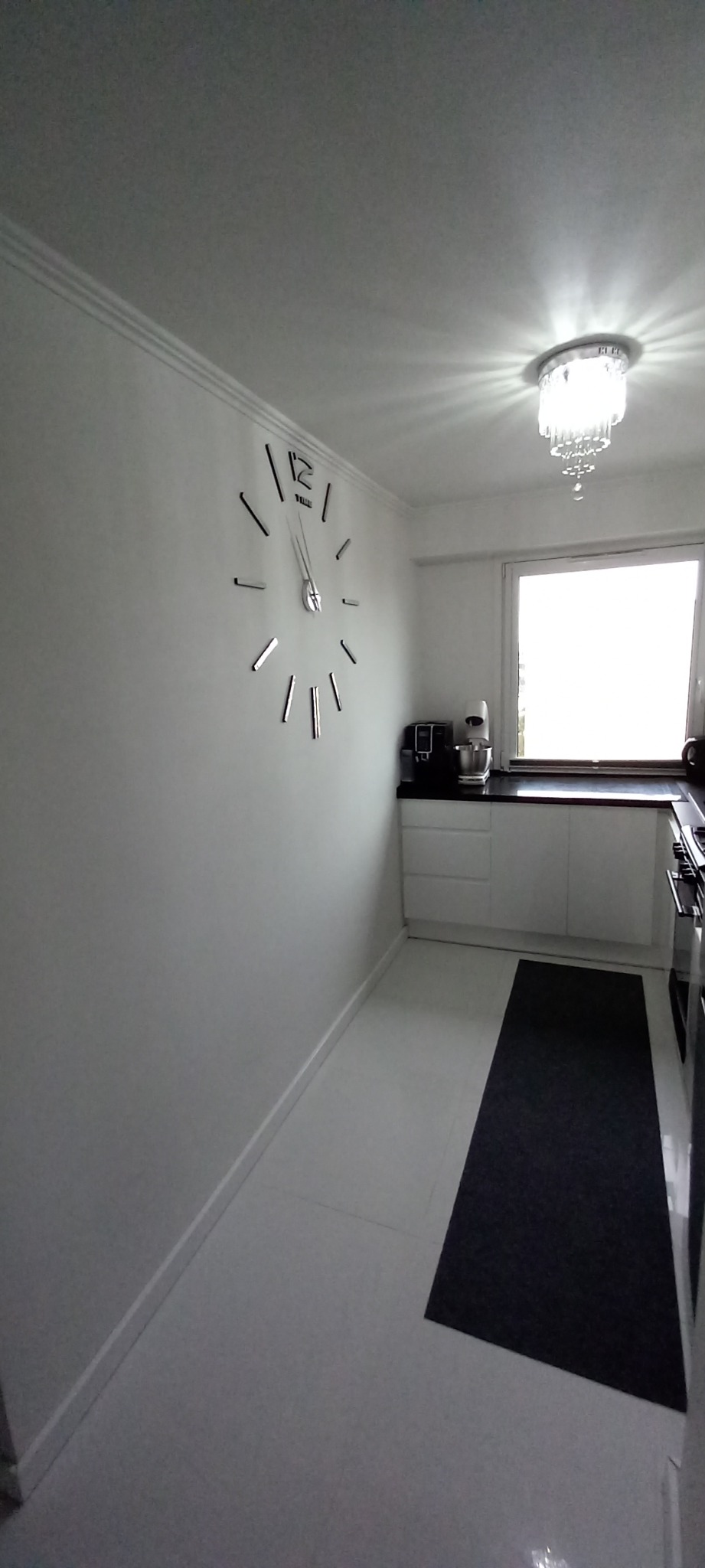 Minimalistyczna kuchnia z białymi ścianami, podłogą i meblami, kontrastującymi z czarnym blatem i dywanem, oświetlona kryształowym żyrandolem, z dużym zegarem ściennym.