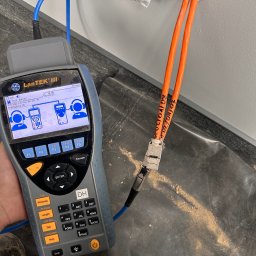 Power Audit - Pomiar kabli sieciowych testerem LanTEK III przy gnieździe w ścianie. Dwa pomarańczowe kable z oznaczeniami. Tester w dłoni, widoczny ekran i klawiatura.