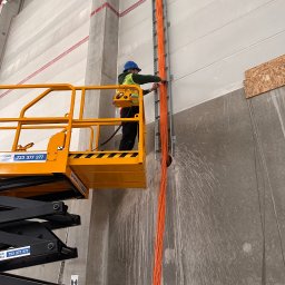 Power Audit - Elektryk na podnośniku montuje pomarańczowe kable w korytku na ścianie hali. Widoczne ślady po wierceniu. Pracownik w kasku i kamizelce.