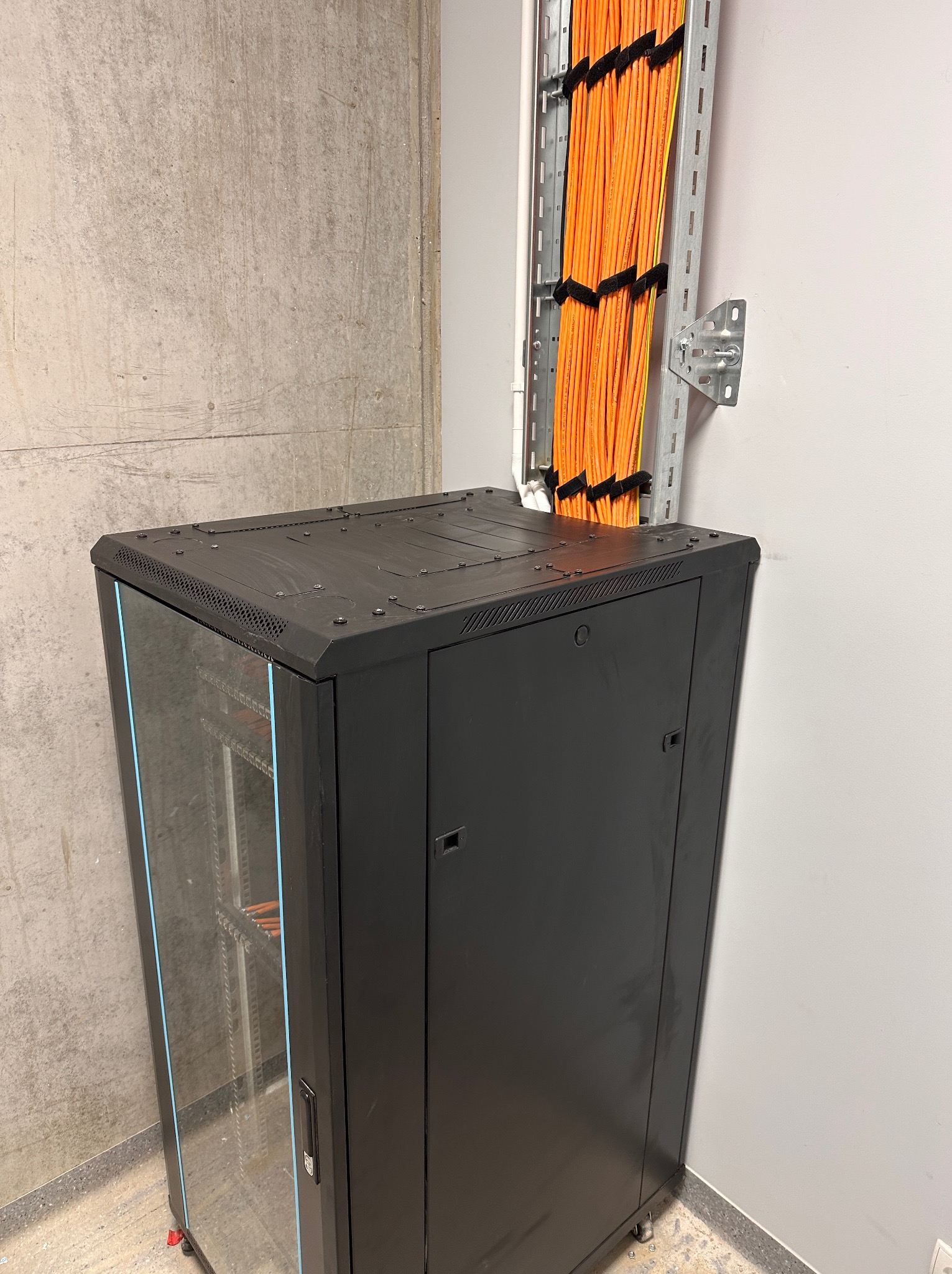 Szafa rack z pomarańczowymi kablami w korycie na ścianie. Widoczne kable wewnątrz szafy przez przeszklone drzwi. Szara ściana i betonowa kolumna w tle.