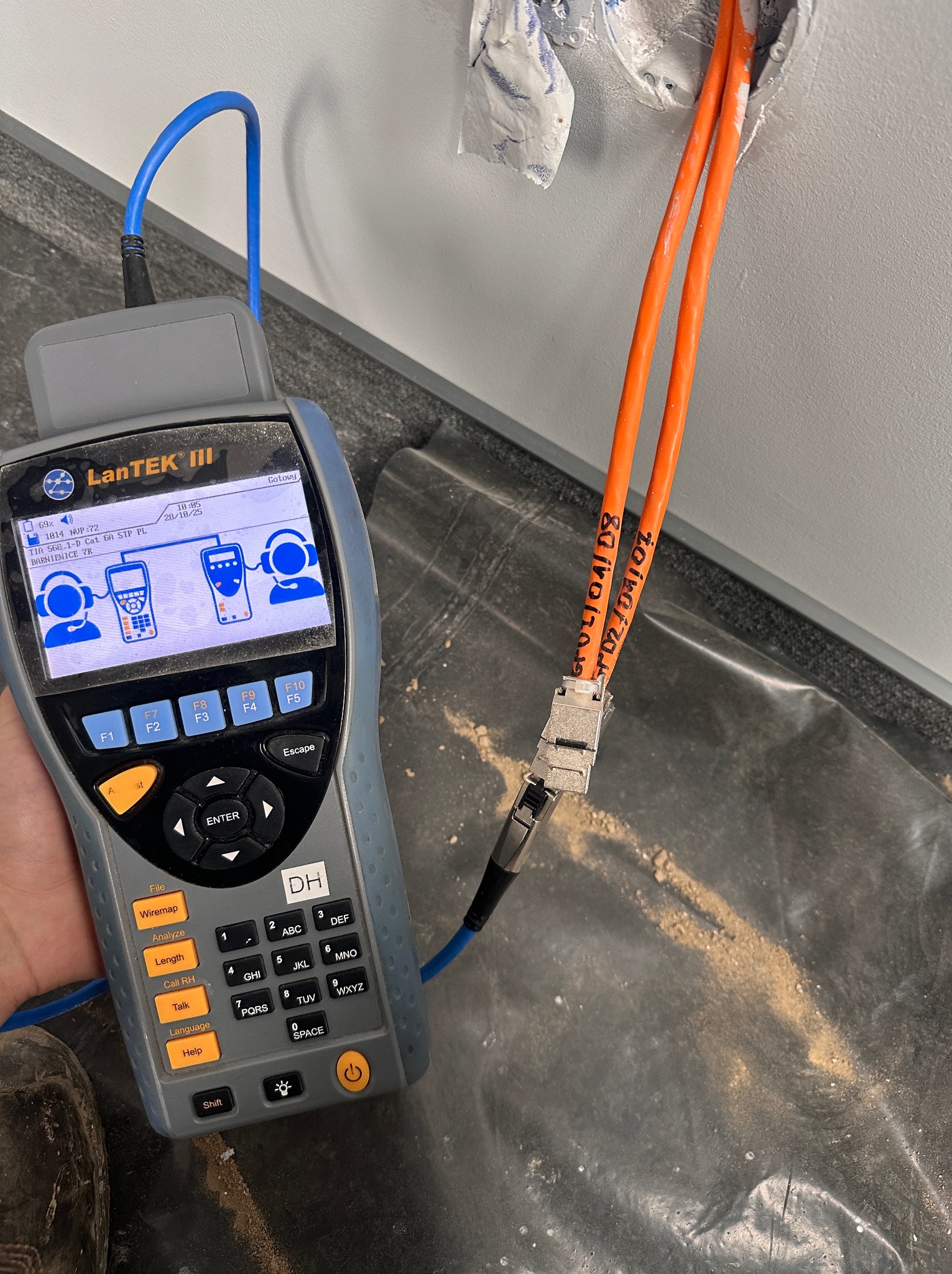 Pomiar kabli sieciowych testerem LanTEK III przy gnieździe w ścianie. Dwa pomarańczowe kable z oznaczeniami. Tester w dłoni, widoczny ekran i klawiatura.