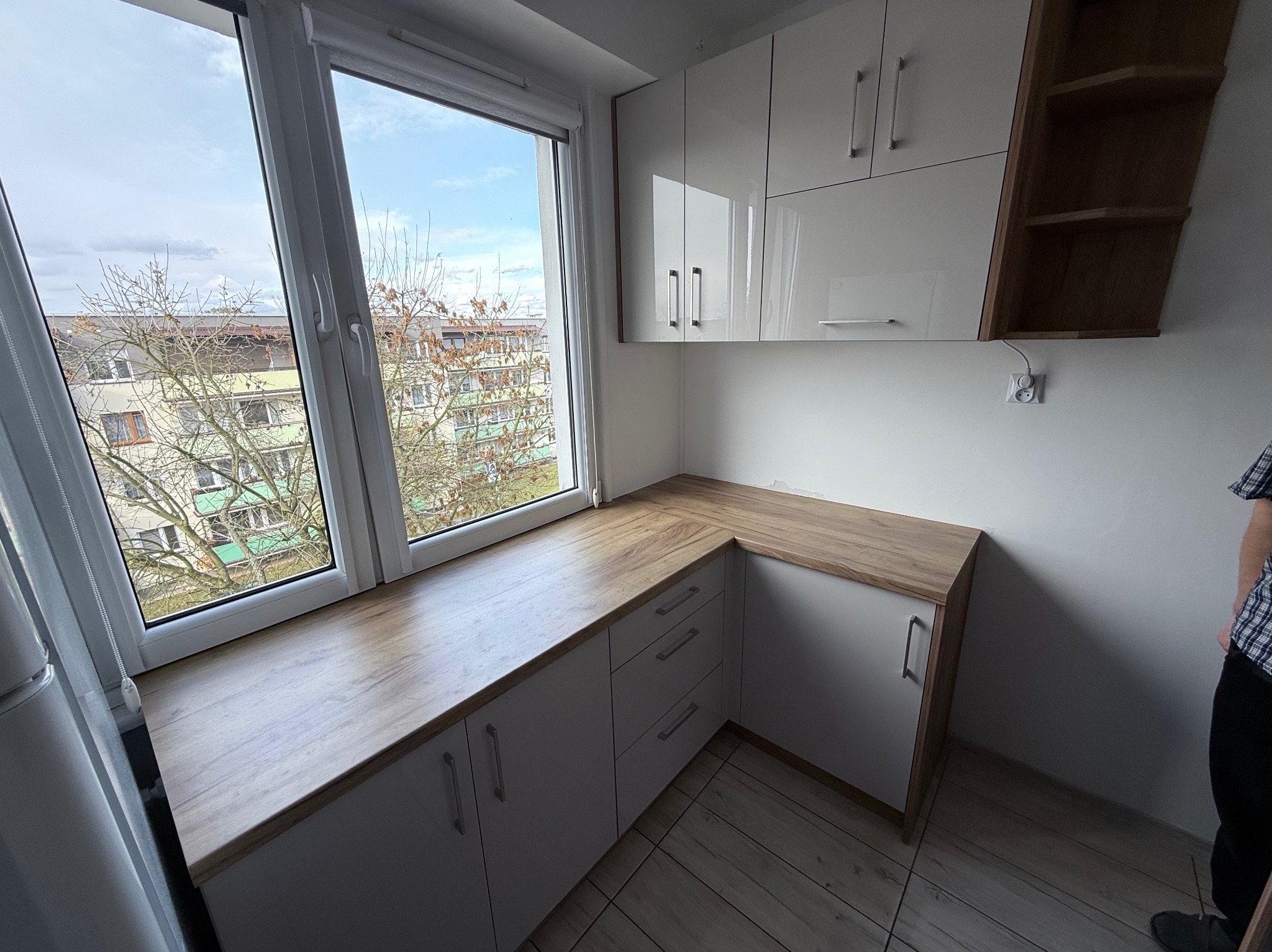 Białe szafki kuchenne pod oknem z drewnianym blatem, widok na miasto. Górne szafki lakierowane na wysoki połysk, obok półki z ciemnego drewna.