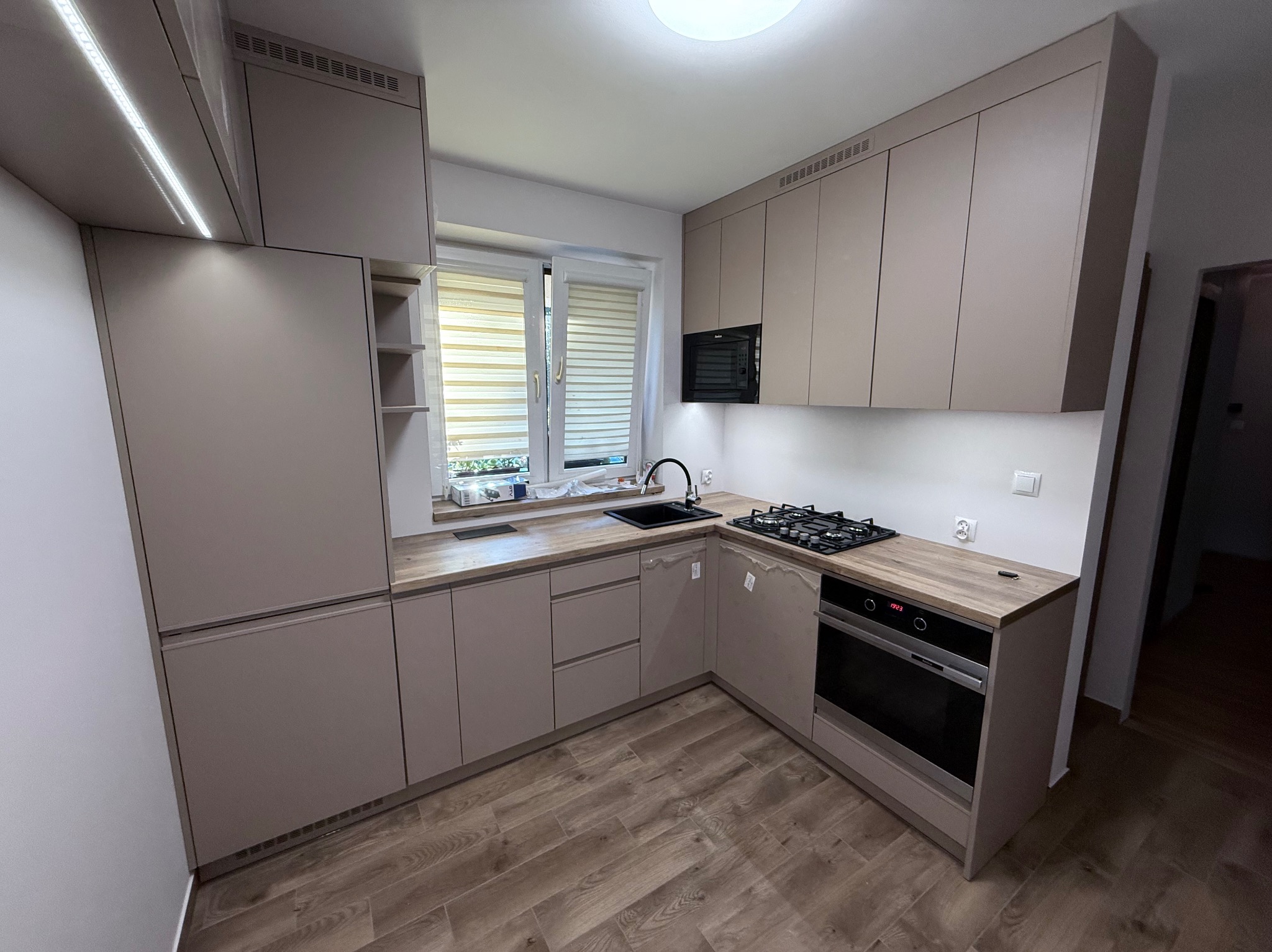 Jasna kuchnia z drewnianym blatem i zabudową pod sufit. Nowoczesny design z oknem i sprzętem AGD. Uwagę przykuwa minimalistyczny styl i funkcjonalność.