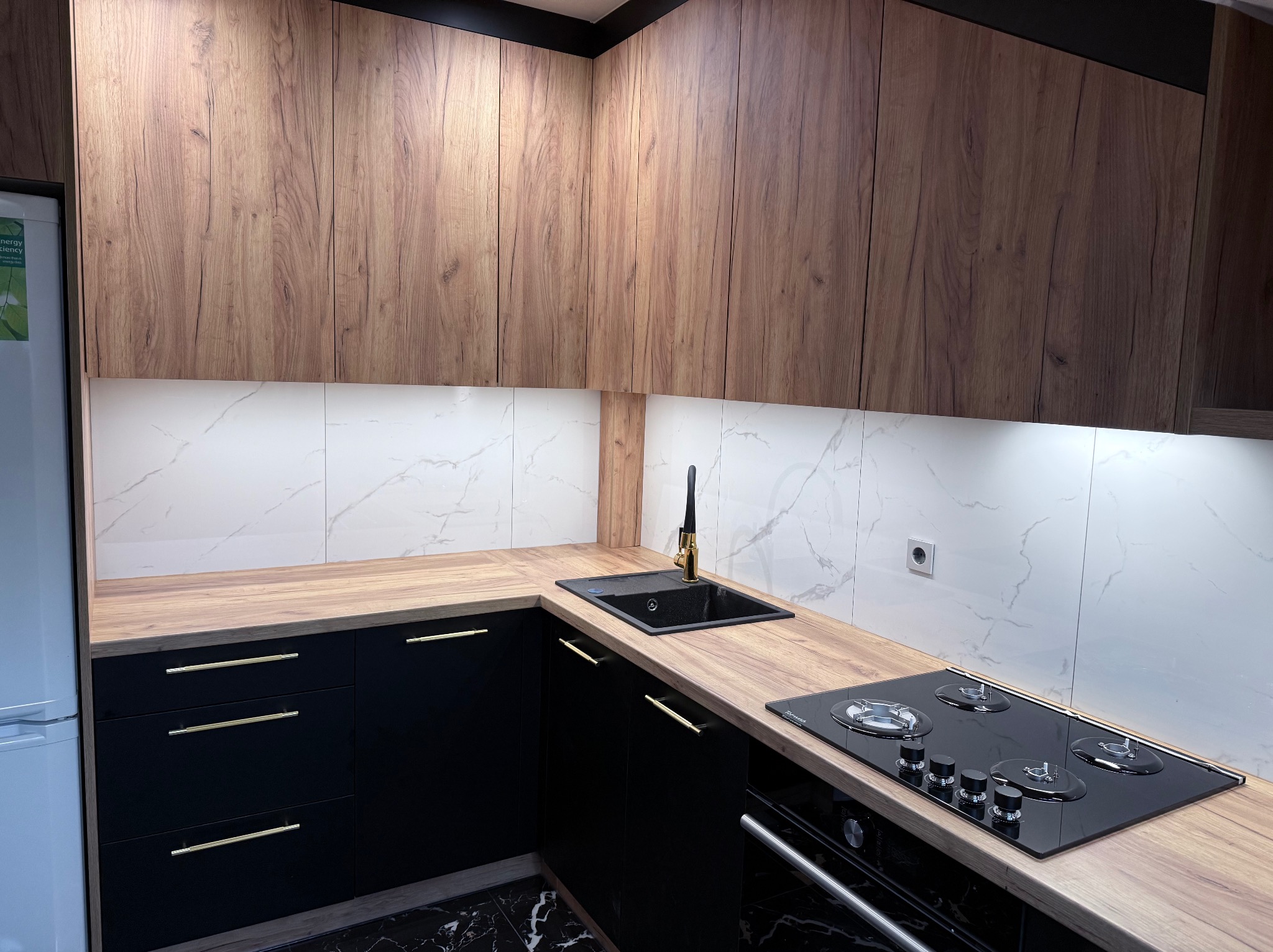Nowoczesna kuchnia z drewnianymi szafkami górnymi i czarnymi dolnymi, złote uchwyty, marmurowe płytki, czarny zlew i płyta gazowa. Oświetlenie LED pod szafkami.