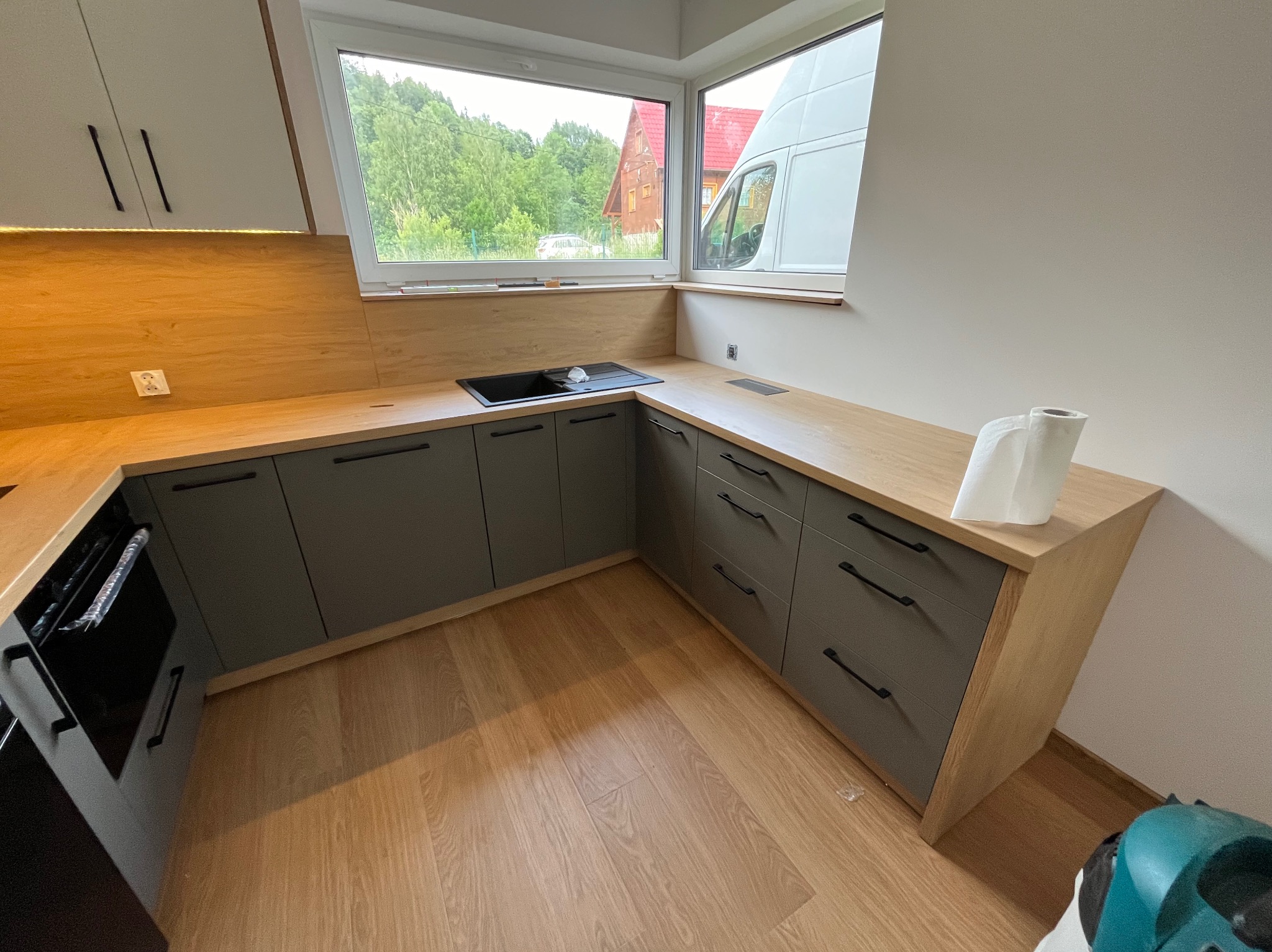 Nowoczesna kuchnia z szarymi szafkami i drewnianym blatem, zlewem pod oknem i widokiem na zieleń. Minimalistyczne uchwyty, jasne drewno i naturalne światło.