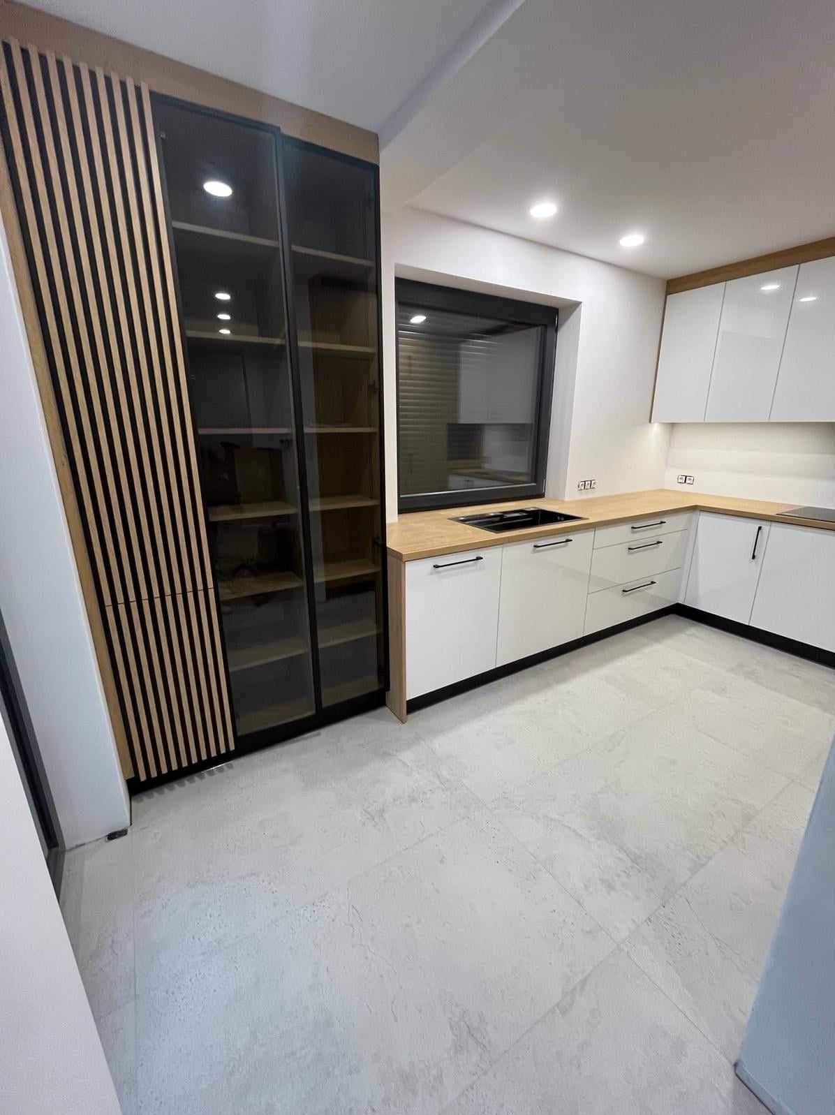 Nowoczesna kuchnia z białymi szafkami, drewnianym blatem i wysoką szafą z lamelami. Minimalistyczny design, jasne wnętrze, czarne akcenty.